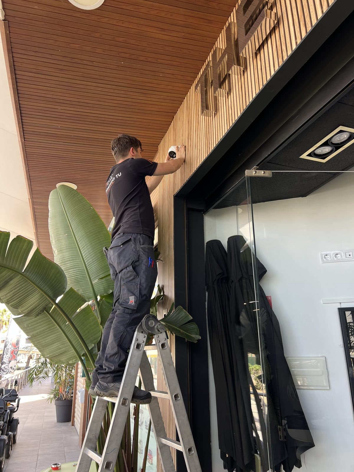 A Buzz Alarmas engineer on a ladder fitting a security camera above a shop entrance.