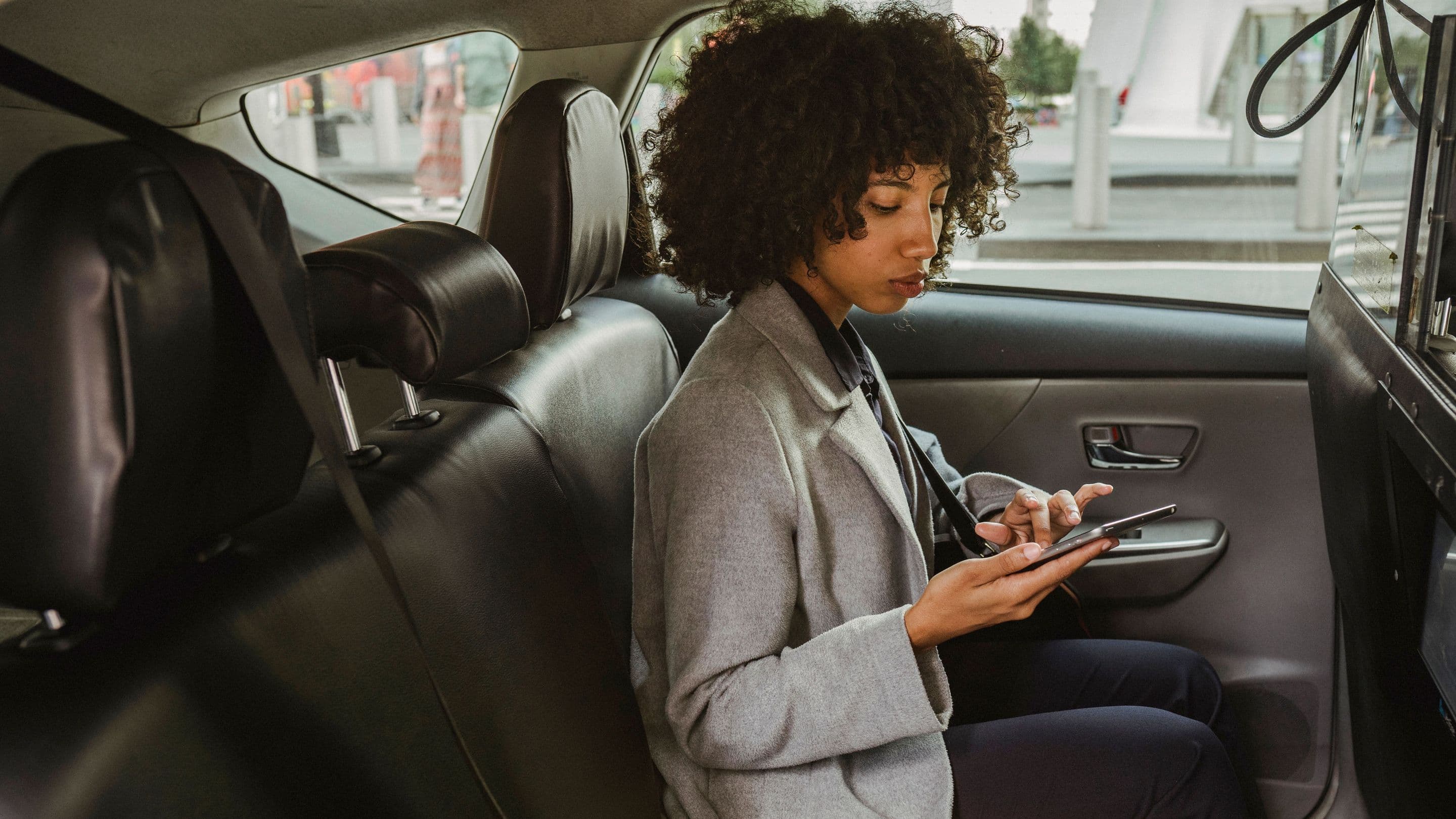 A woman in a grey coat in the back seat of a car, typing on her phone.
