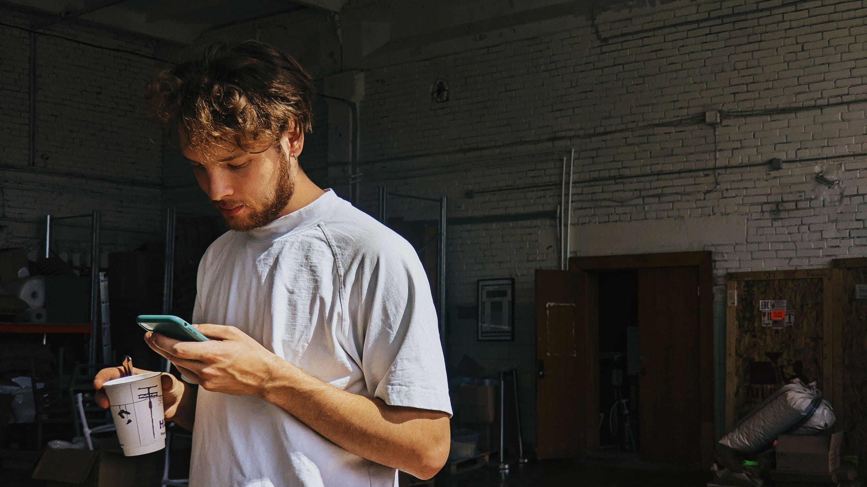 A man in a white T-shirt in a brick workshop, reading his phone, a paper cup in his other hand.