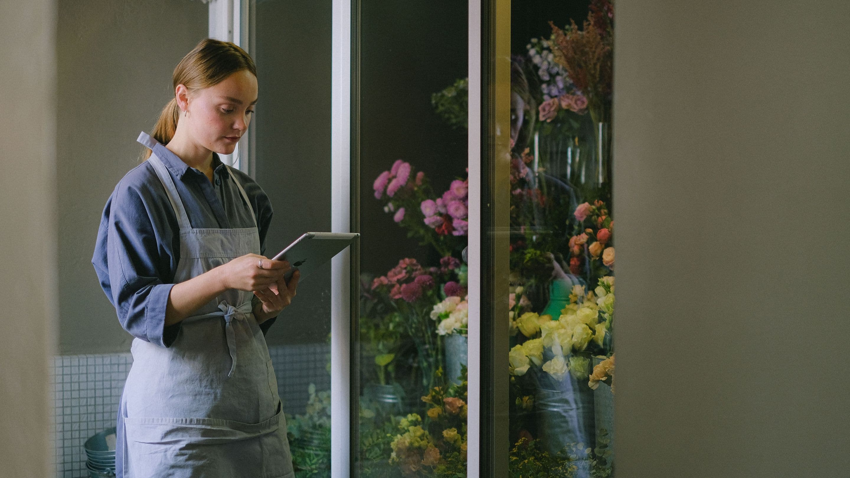 A florist in an apron in her shop, reading a tablet beside the flower fridge.