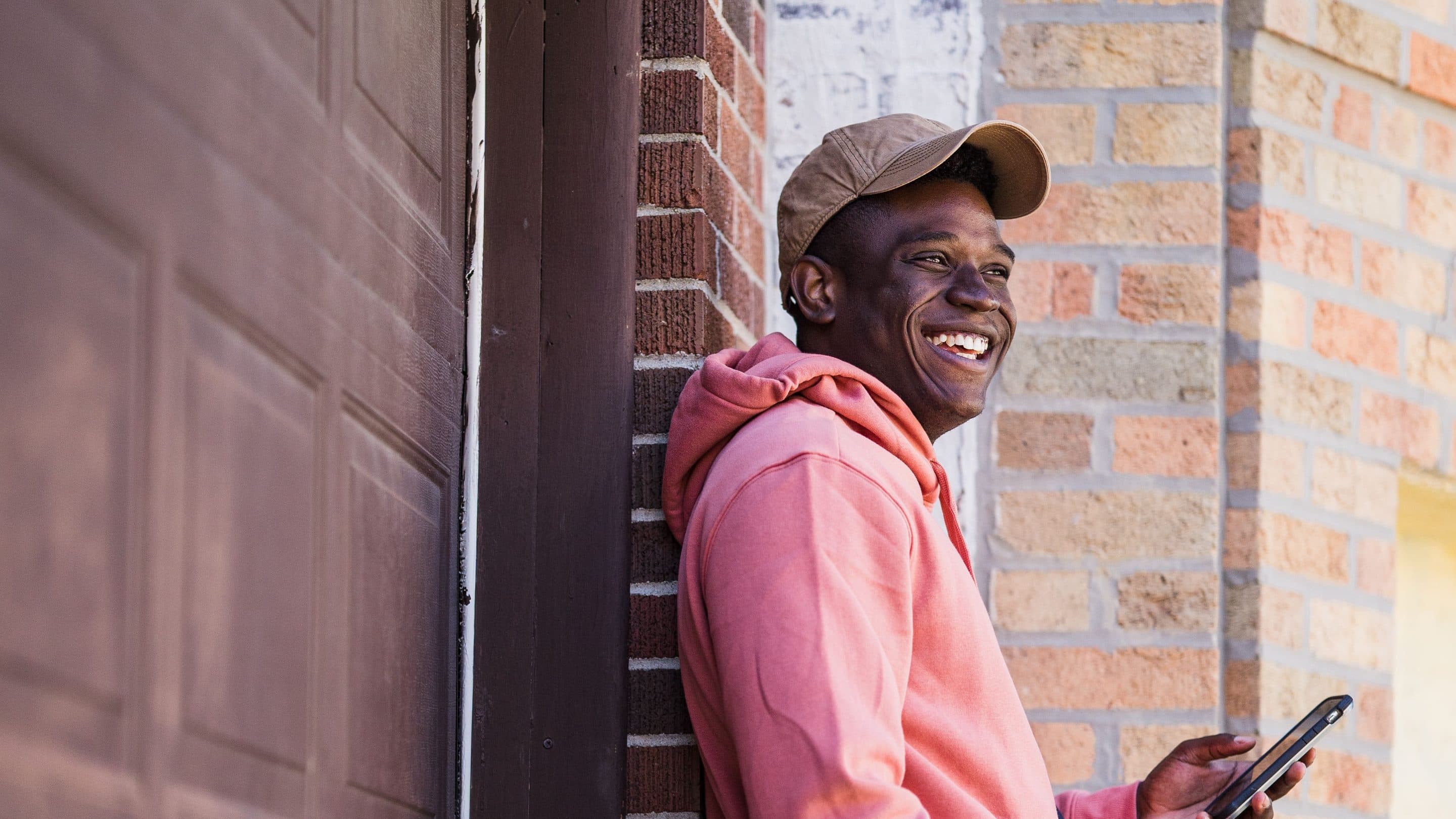 A man in a pink hoodie leaning on a brick wall, laughing, phone in hand.